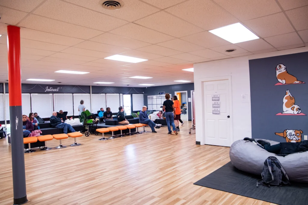 Large activity room with adults, staff, seating areas, and open space at an adult day support program.