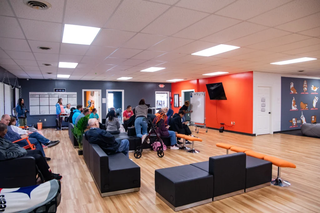 Large activity room with adults and staff gathered for a group session at an adult day support program.