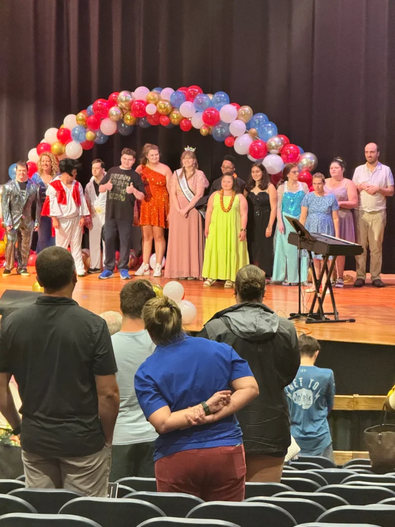 Adults standing on stage under a balloon arch during a community event with an adult day support program.