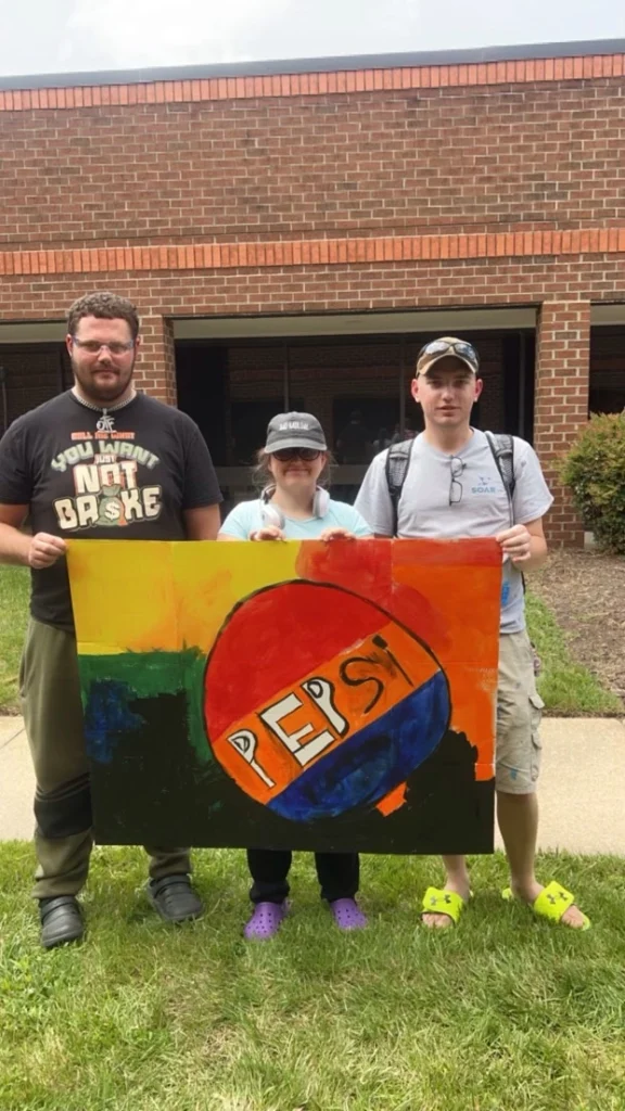Adults standing outside and holding a colorful Pepsi artwork during an adult day support program activity.
