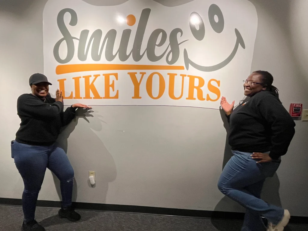 Two Smiles Like Yours staff members smiling and posing beside the program logo wall.