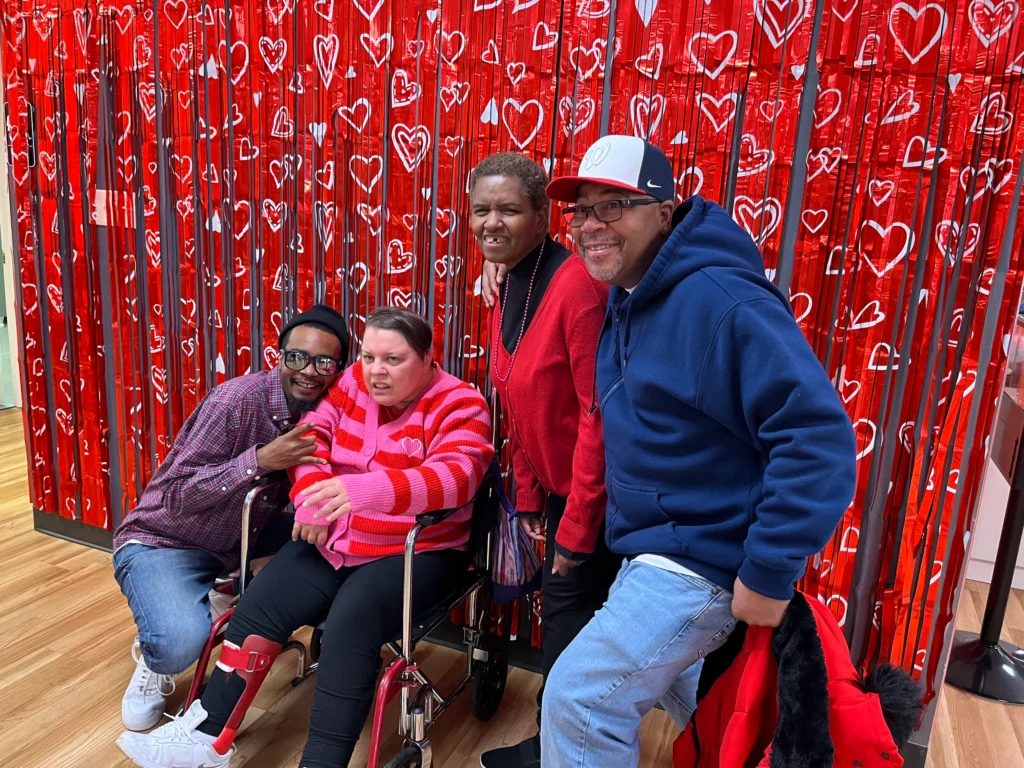 Adults posing together in front of a red heart backdrop during an adult day support program event.