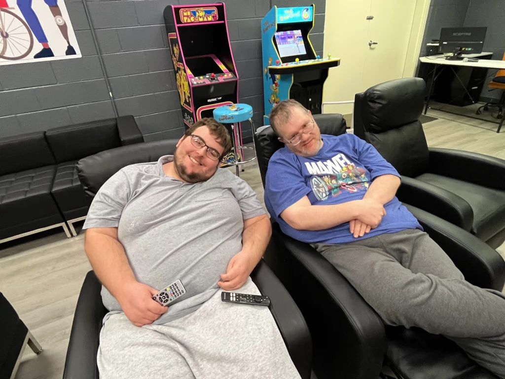 Two adult members relaxing in lounge chairs inside a recreation room at an adult day support program.