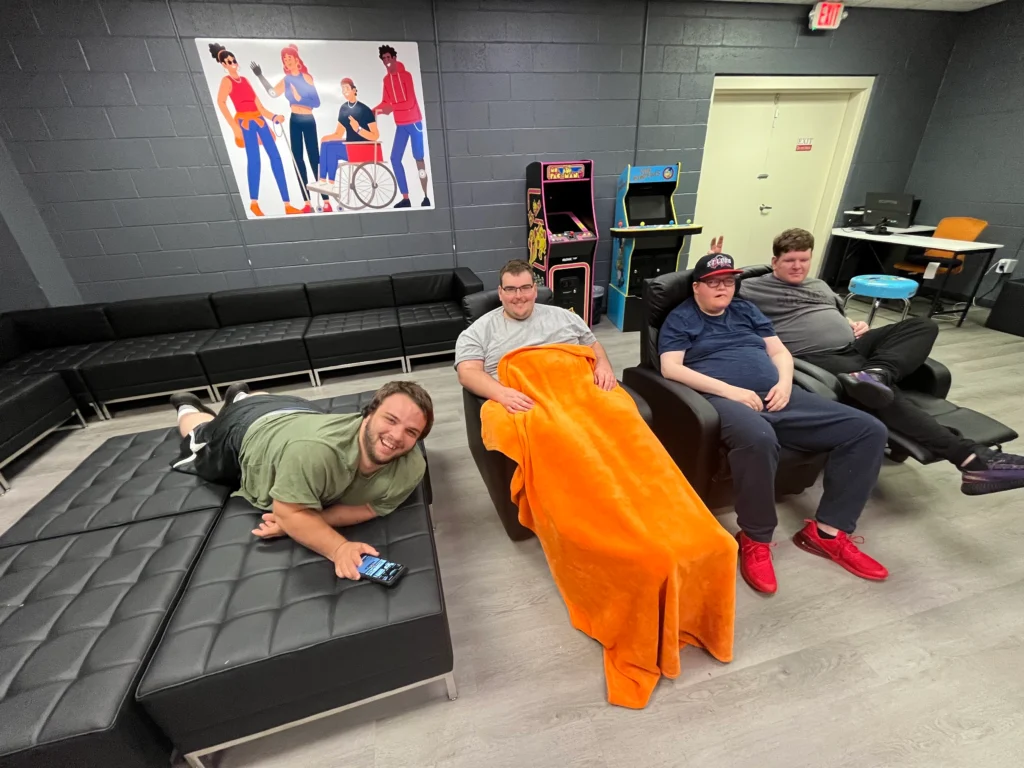 Adults relaxing in a recreation room with lounge chairs and arcade games at an adult day support program.