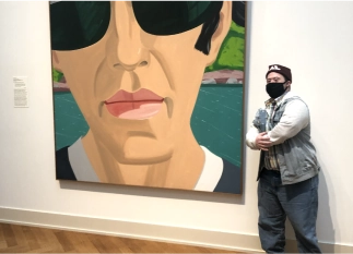 Person standing beside a large artwork of a face wearing sunglasses, showcasing engagement with art in a gallery setting.