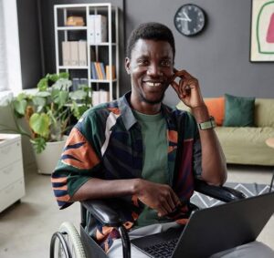 Man using a wheelchair smiling while working on a laptop in a comfortable home setting.