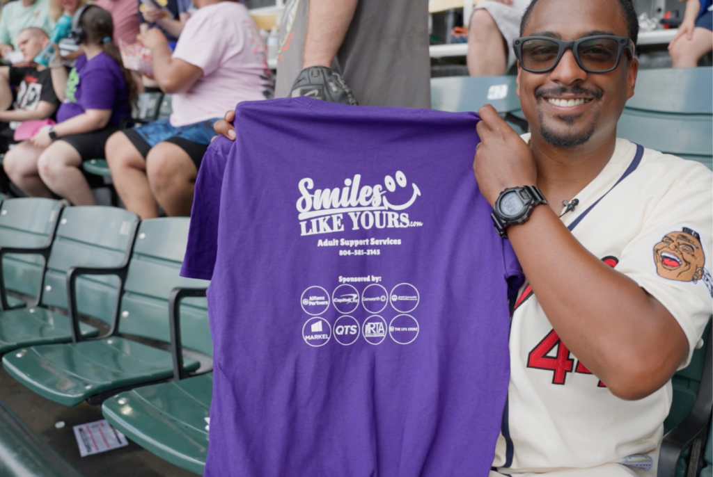 Smiling man holding a purple Smiles Like Yours Adult Support Services shirt at a baseball game.